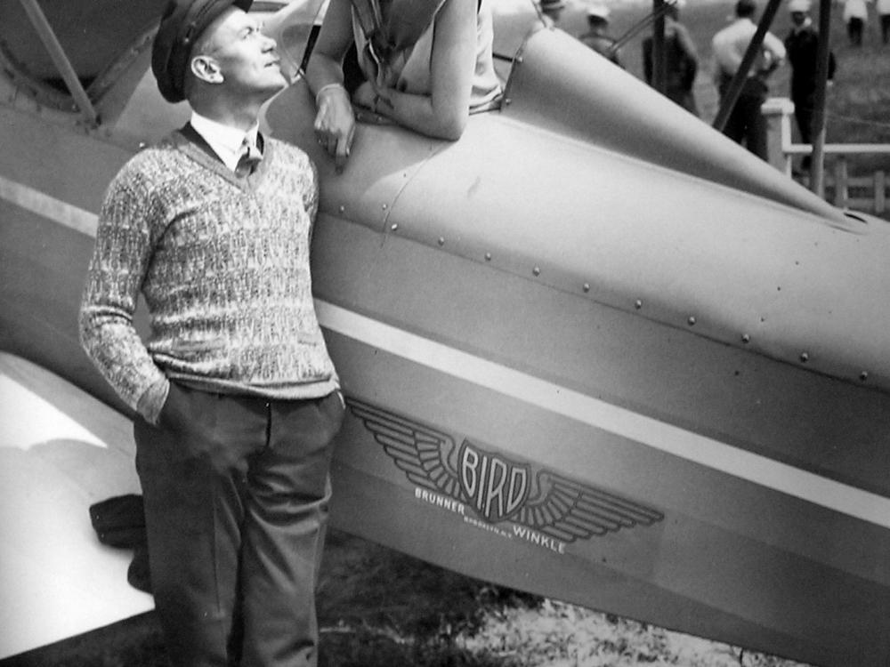 A woman in a pilot's cap leans out of a plane. A man standing on the ground stares up at her. The photo is in black and white.