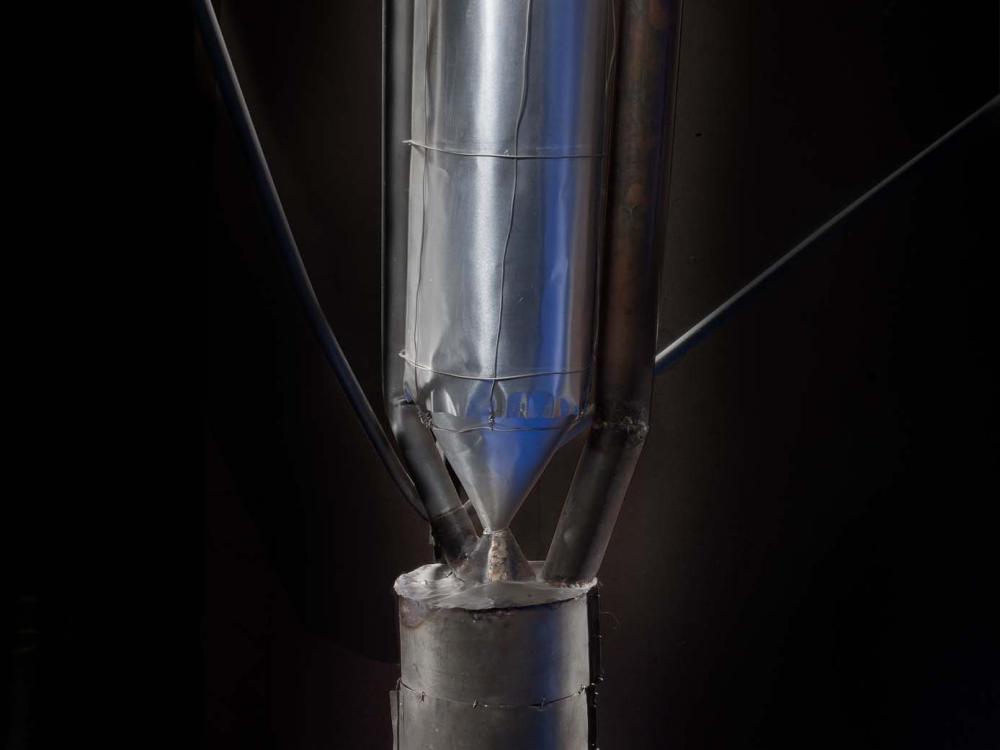 Close-up view of motor on a primitive rocket. A conical nose tops a metal cylinder-shaped motor attached to a cylinder-shaped fuel tank by a metal conical-shaped drain.