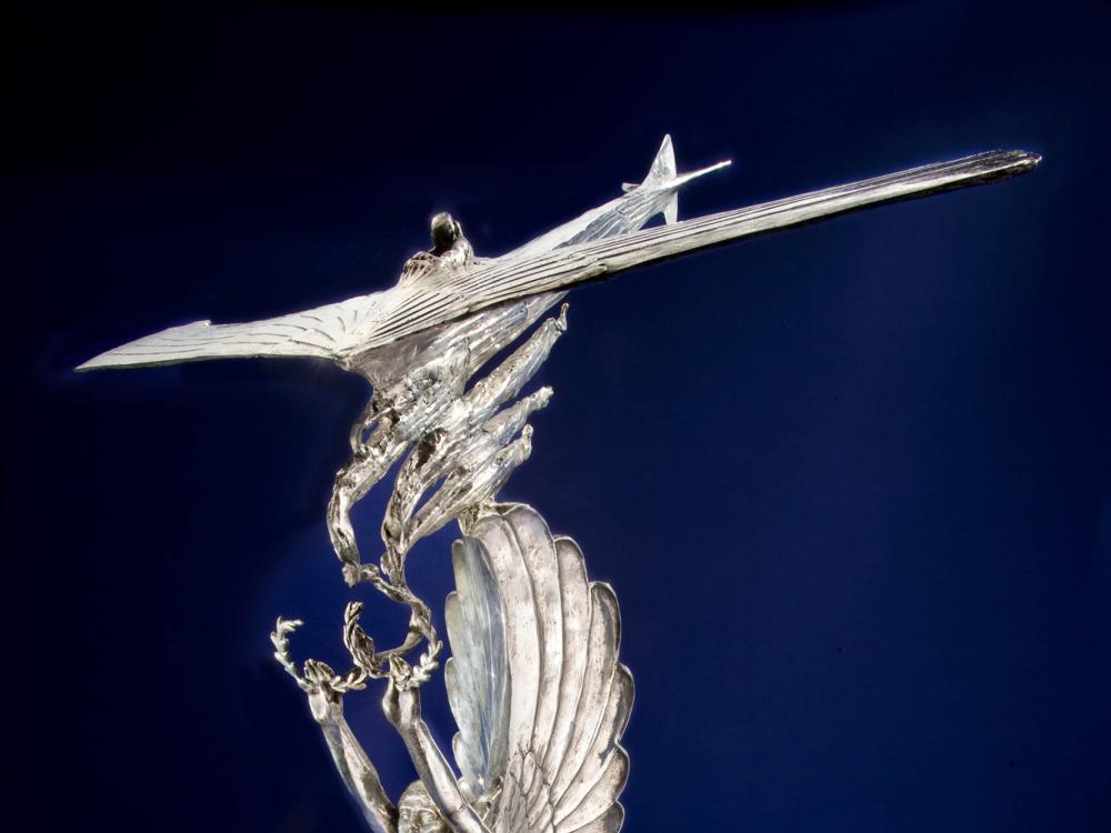 Silver-colored trophy with topper of a winged angel holding olive wreaths which are picked up by a plane.