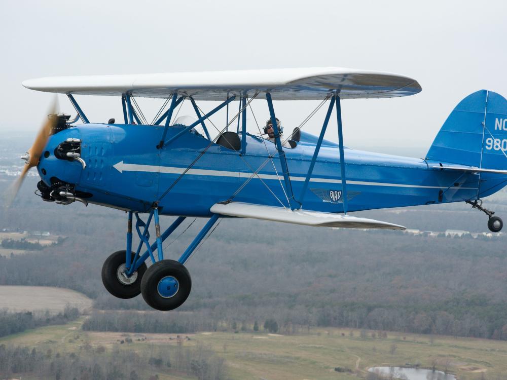 Side view of blue biplane with white wings, one engine, and fixed landing gear. Biplane is in flight. Registration number "N980V" painted in white paint on rudder.