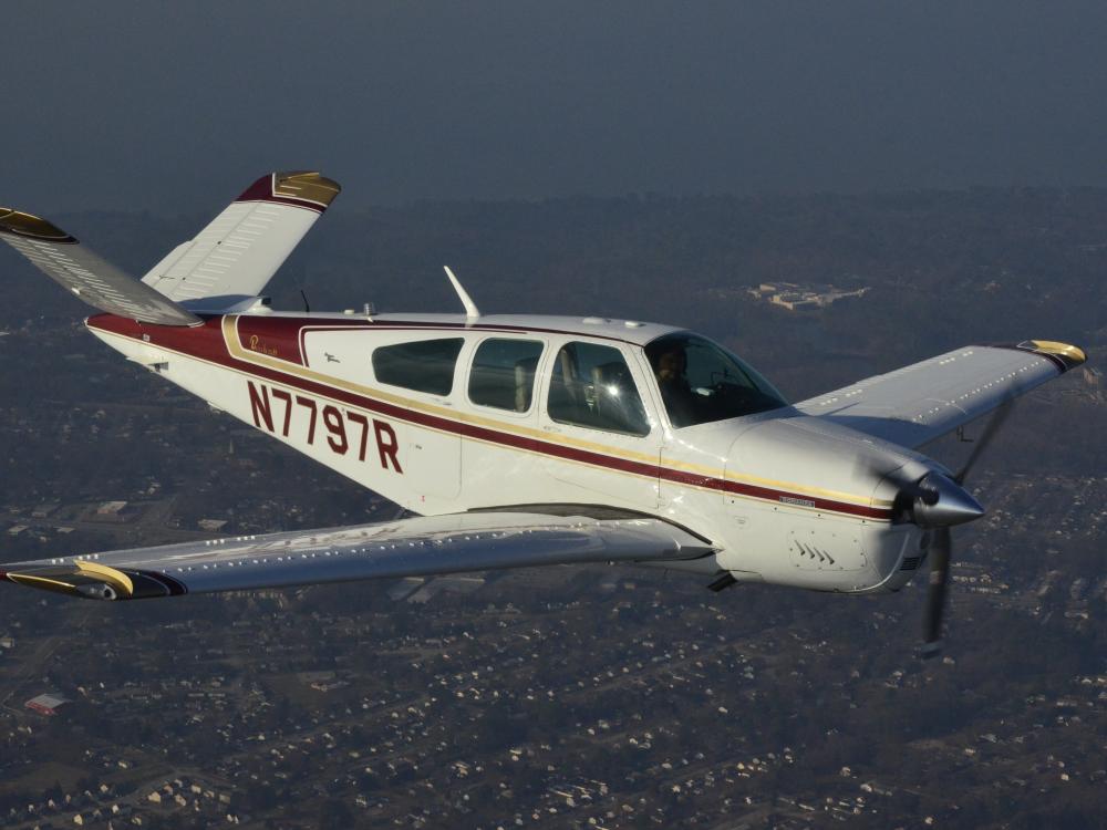 Side view of white and red monoplane in flight. Monoplane has one engine and registration number "N7797R" written in red on the back half of the fuselage.