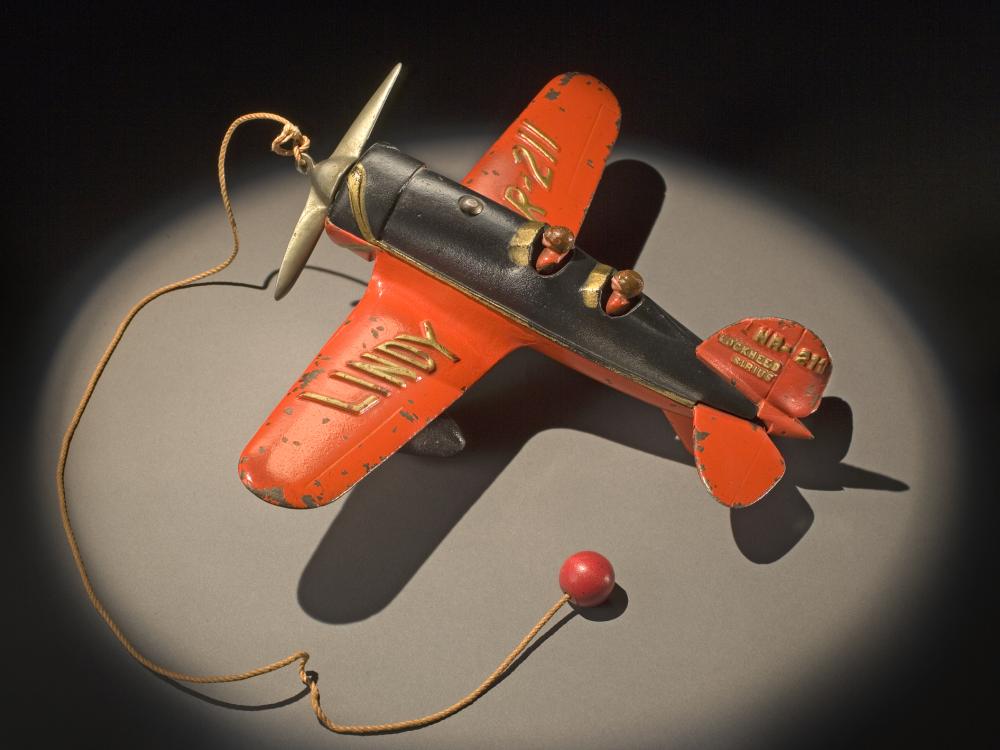 Top view of orange and black monoplane toy with a ball and string attached to the center of the propellors on the front.