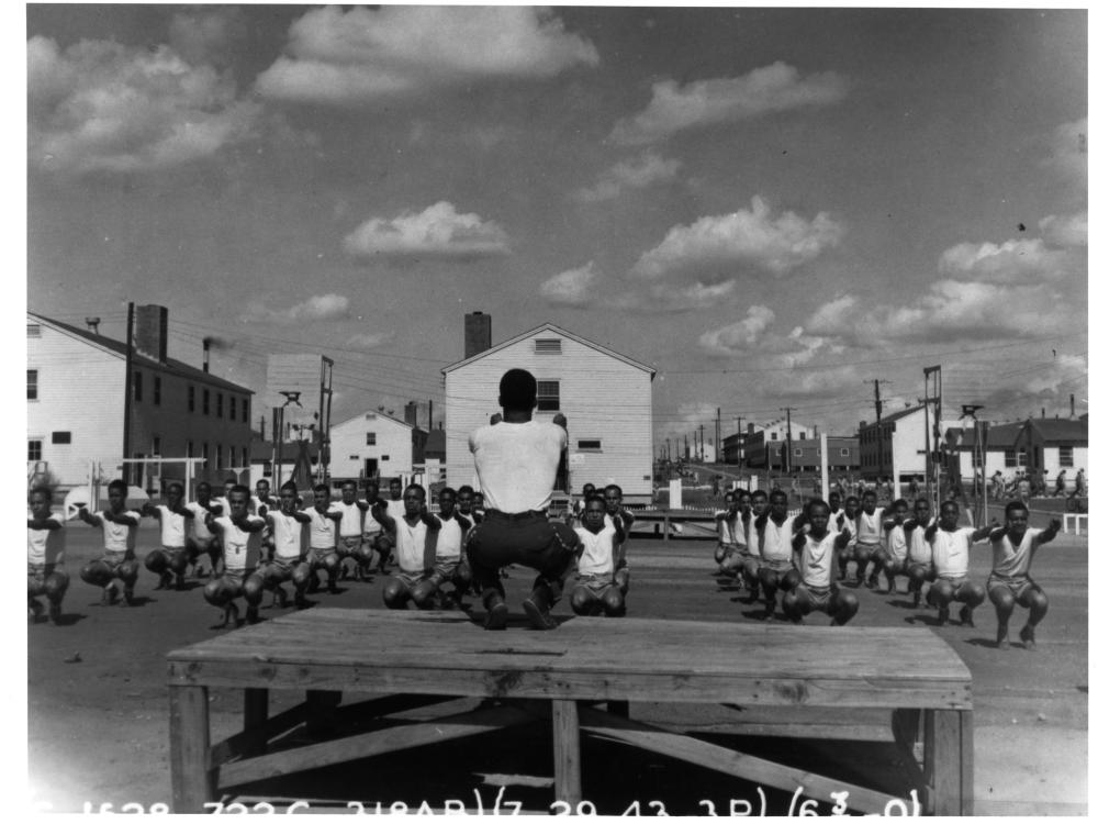 Partial view of some of the buildings and a clearing at the Tuskegee Army Air Field. A person runs physical training for cadets in the foreground.