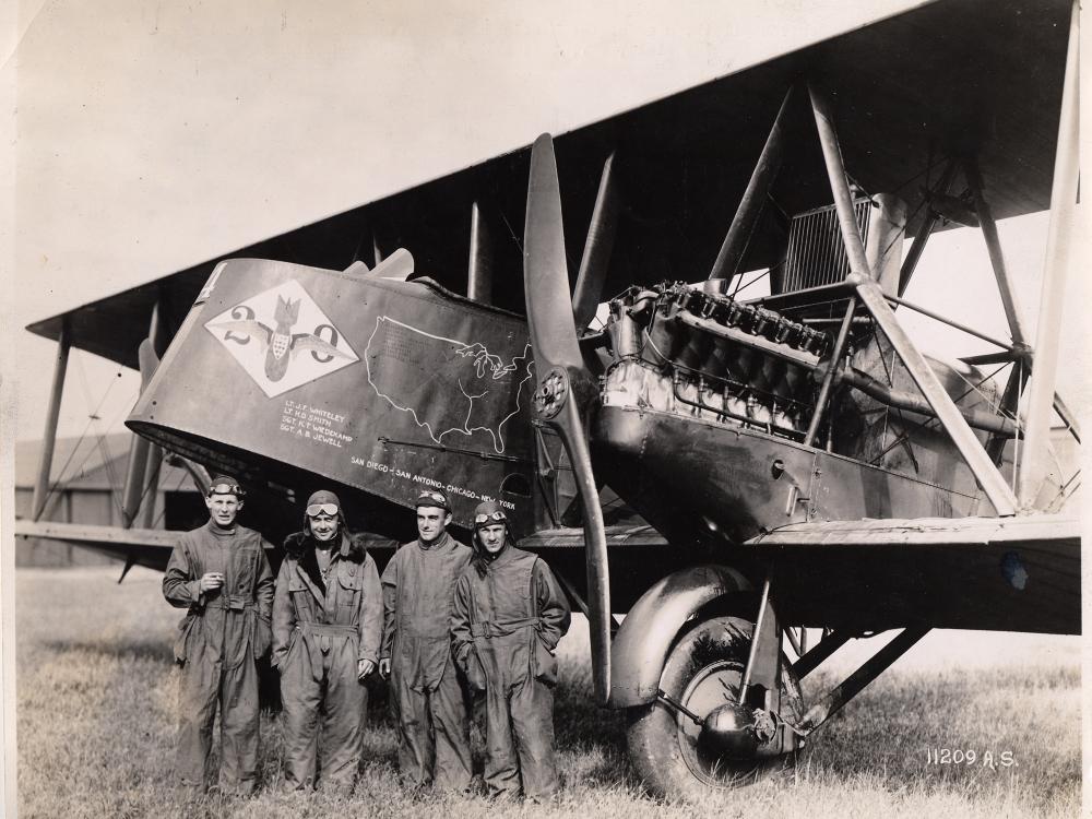 A group of white males representing the U.S. Army Air Service stand next to the nose of a biplane.