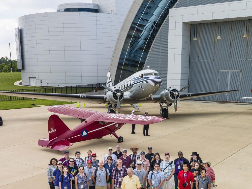 A group photo featuring several social media users who follow the Museum, the volunteers of an event, and the Museum's former director. They are standing on the runway in front of two aircraft.