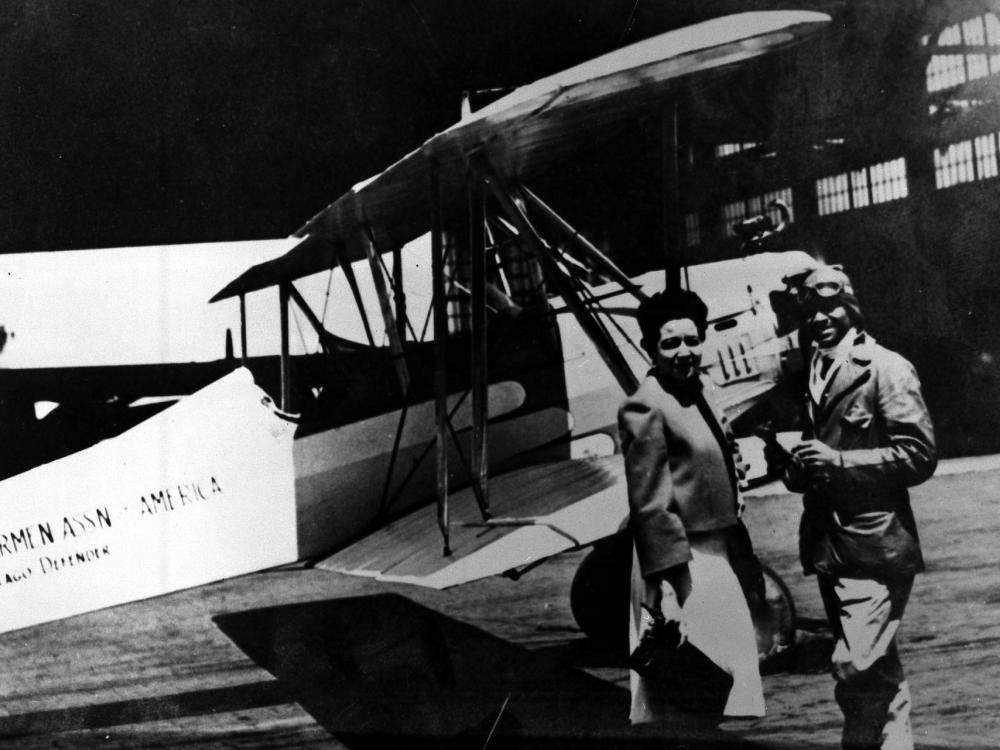 Chauncey Spencer, an African-American male pilot, stands next to a biplane with another person.