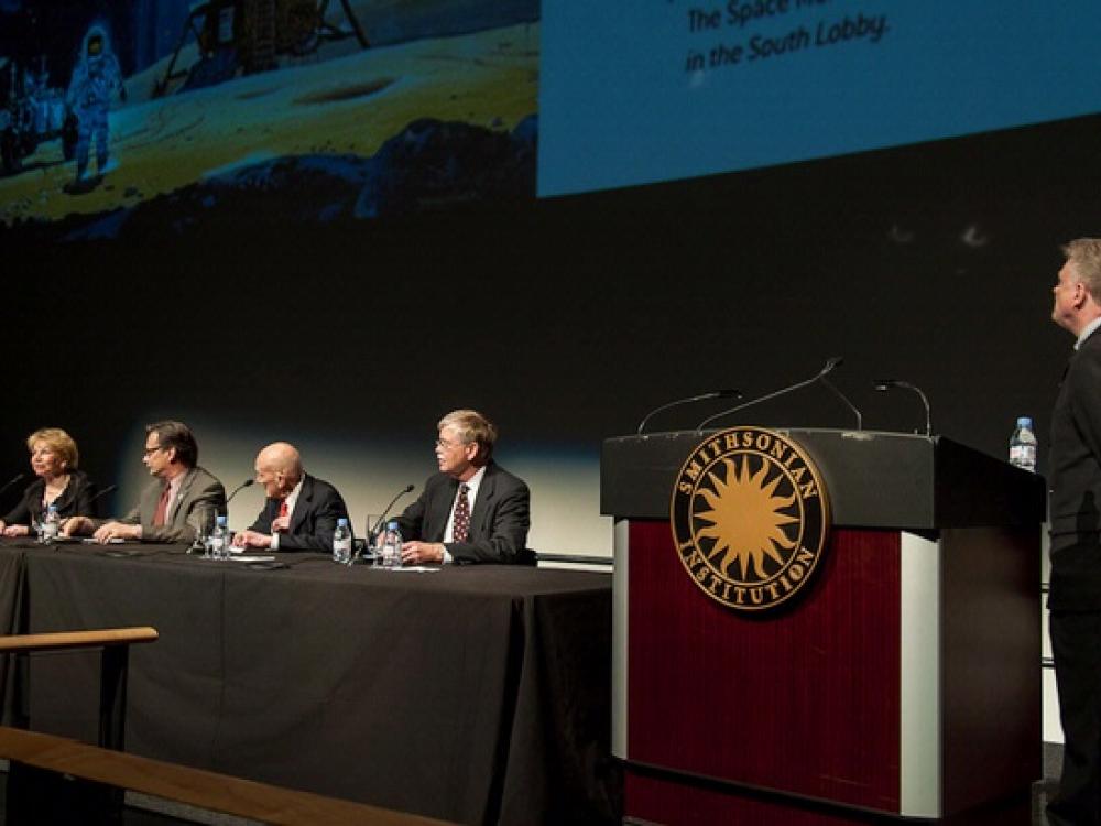 Five people present a lecture at the Museum. One is standing next to a podium whereas four other people are sitting at a table.