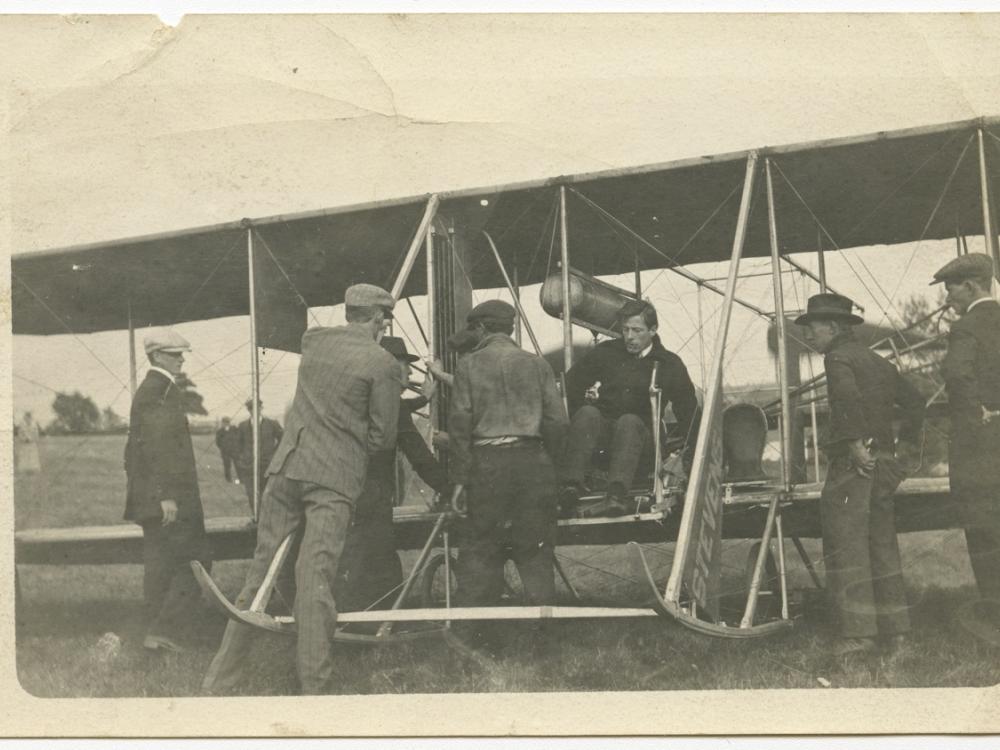 Harry Bingham Brown, a white male pilot, prepares for takeoff in his Wright biplane with the help of several other men.