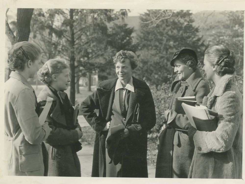 Amelia Earhart, a white female pilot, stands in the center of a group of five people. She is smiling toward the people on the left.