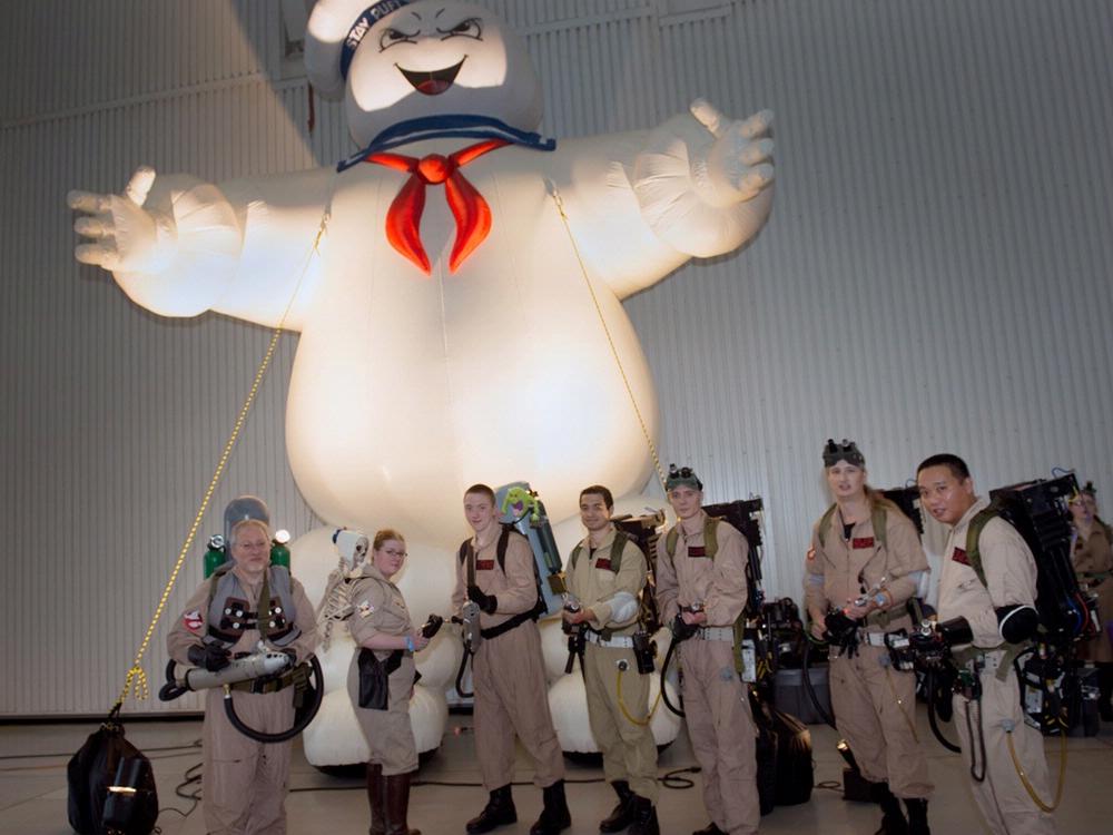 People dressed up in Ghostbusters costumes stand together in front of a Ghostbusters ghost float inside the Museum.