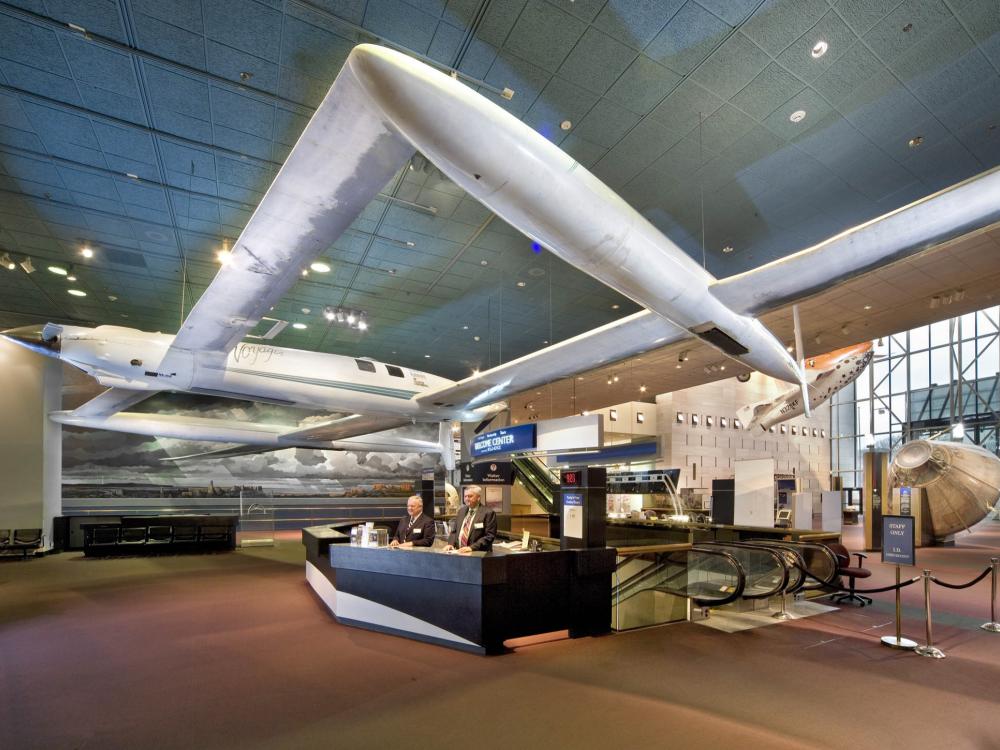 Two docents stand at the museum's welcome desk. A white, twin-boom aircraft hangs above the desk.