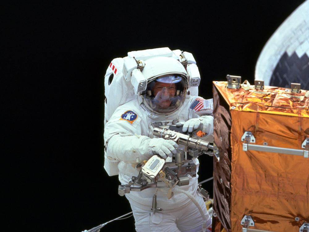 Claude Nicollier, a male European Space Agency astronaut, uses a power tool on an orange-colored storage enclosure during a spacewalk.