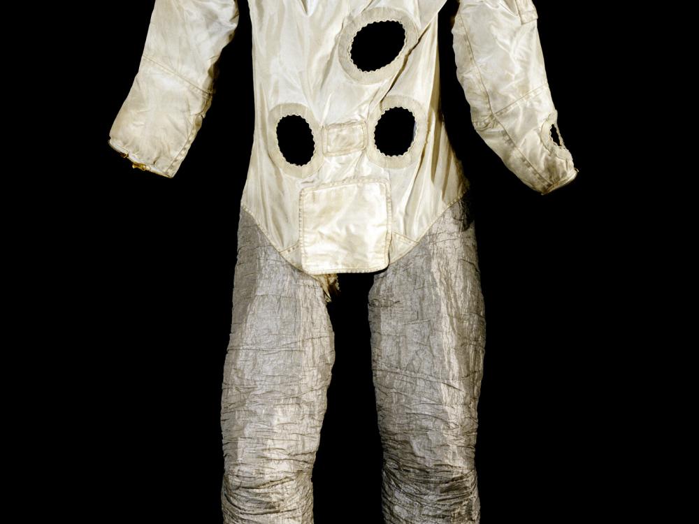 Front view of astronaut suit used for astronaut training on Earth. The top half features a white fabric with multiple holes representing life support tubes, whereas the bottom half features a silver-colored fabric.