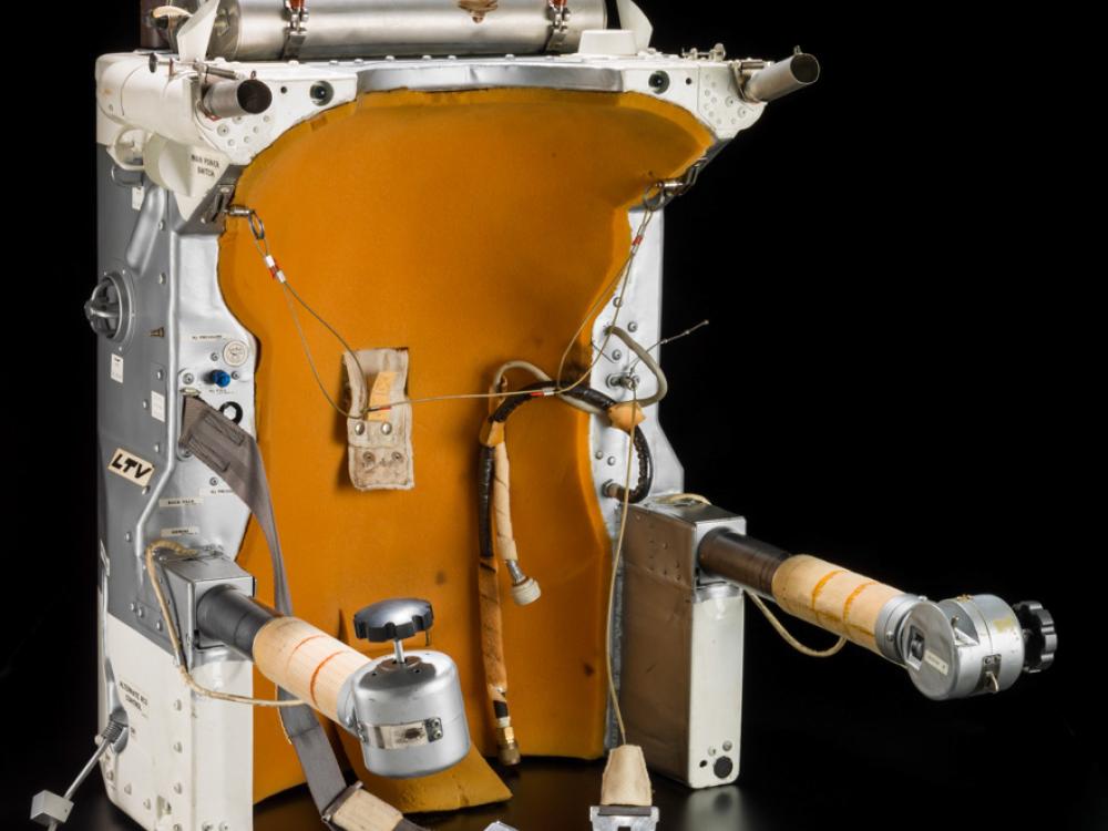 Orange, white, and silver-colored object with silver-colored straps to restrain a human. Arms with buttons at the end of the arm are used to navigate astronauts in space.