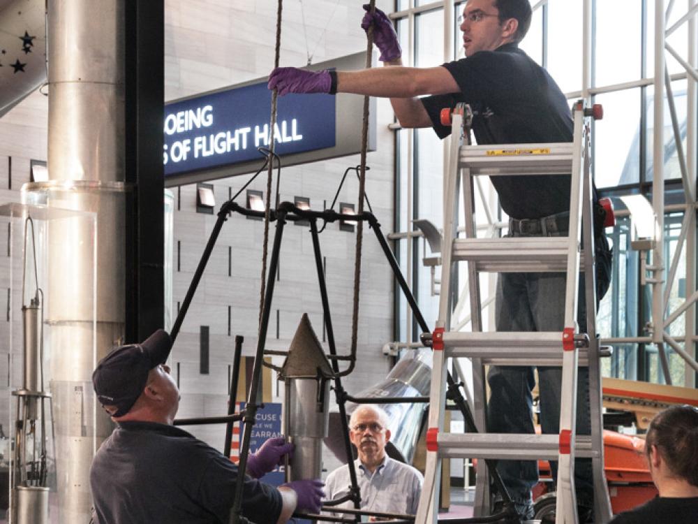 Staff members work to disassemble a primitive rocket from display in the Museum. One person is on a ladder while an other person holds the frame structure together.