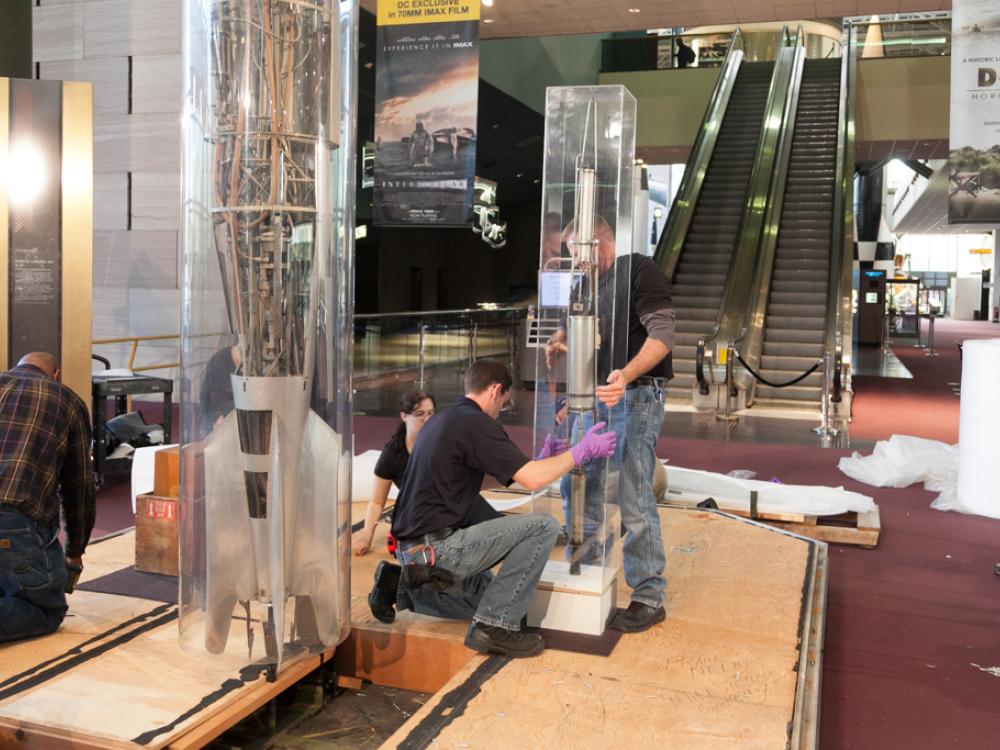 Three Museum staff members work to remove a primitive rocket from a protective case prior to transporting the rocket.