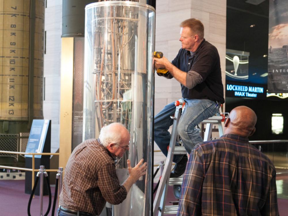 Three Museum staff members work to remove a 1940s rocket from a protective case prior to transporting the rocket.