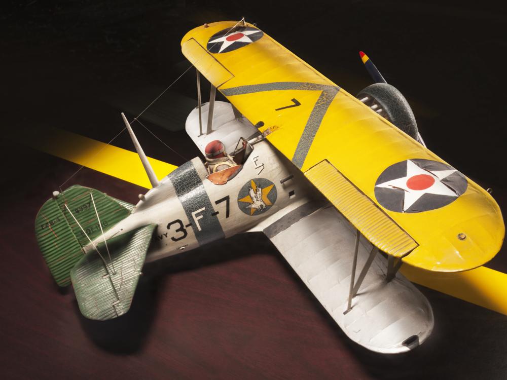 Top sidal view of yellow and silver-colored biplane model aircraft. One white star inside a blue circle is featured on each side of the top wing and identification number "3-F-7" is visible in black paint on the side of the fuselage.