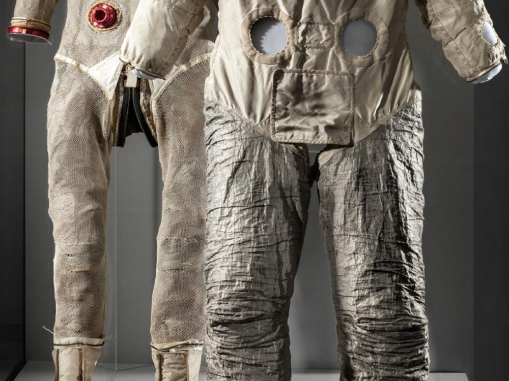 Two suits used by one astronaut on display at the Museum. One is a tan brown spacesuit without helmet and gloves, but the life support tubes are present. The second suit is a tan brown training suit used on Earth and features the life support tubes missing alongside the gloves and helmet.