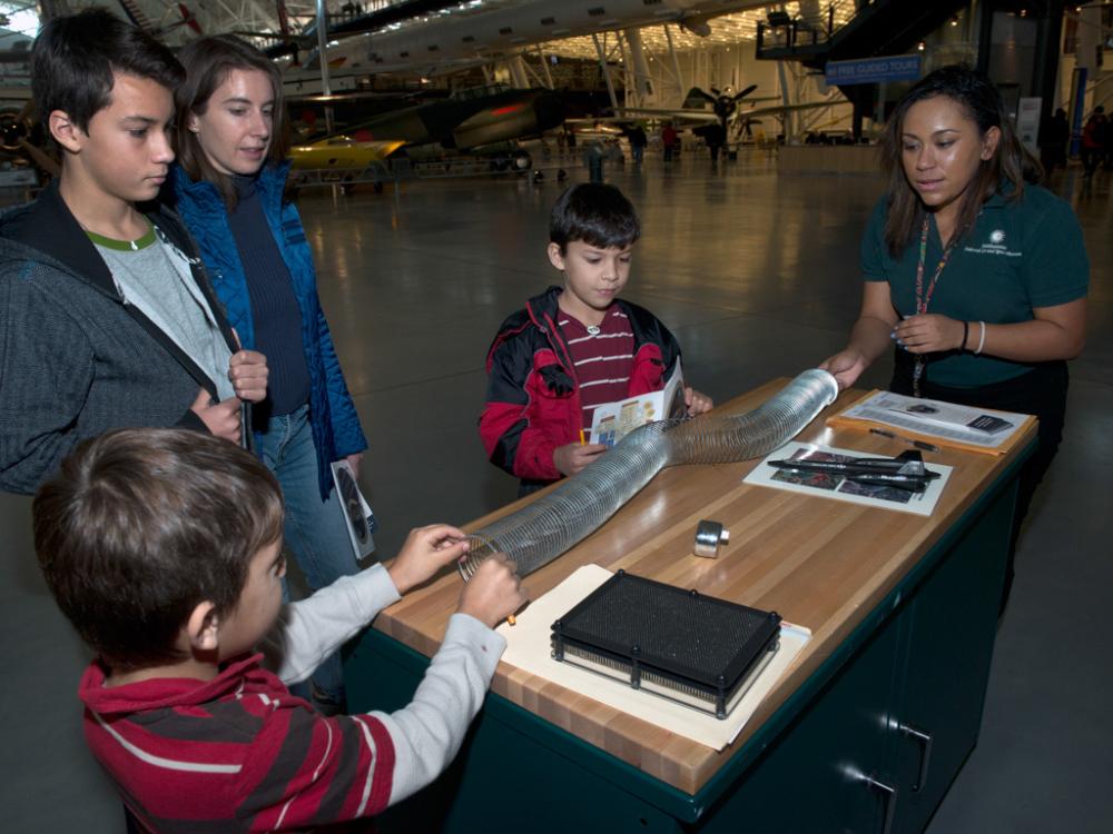 Four Museum visitors and one member of the Museum's staff work together to solve a game. Two visitors and the staff member are playing with a silver-colored twisted object.