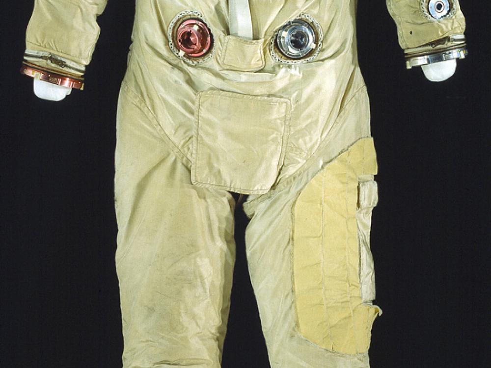 Front view of full-body spacesuit made of tan fabric. Suit does not include gloves or helmet, but it includes the life support tubes. NASA patch on right chest and nametag on left chest.