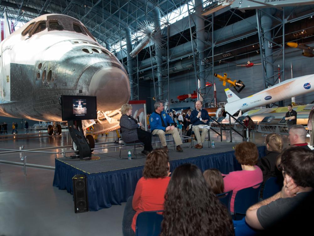 Three Museum staff members host a presentation in front of the Space Shuttle Discovery, a white aircraft designed for space travel, during an open house event.