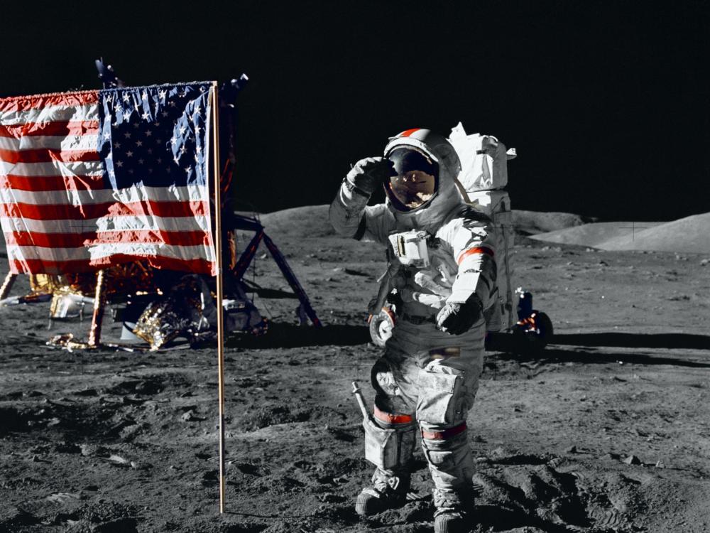 Eugene A. Cernan, a male astronaut wearing a full-body spacesuit, salutes a red, white, and blue U.S. Flag on the surface of the Moon.