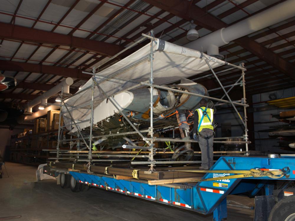 A gray and white metal aircraft, attached to a steel support structure, sits on the long and large bed of a transportation truck. A white tarp protects the top of the aircraft.