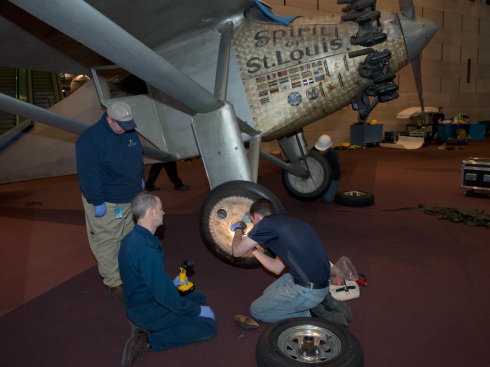 Three Museum staff members work to change tires on the Spirit of St. Louis, a silver monoplane.