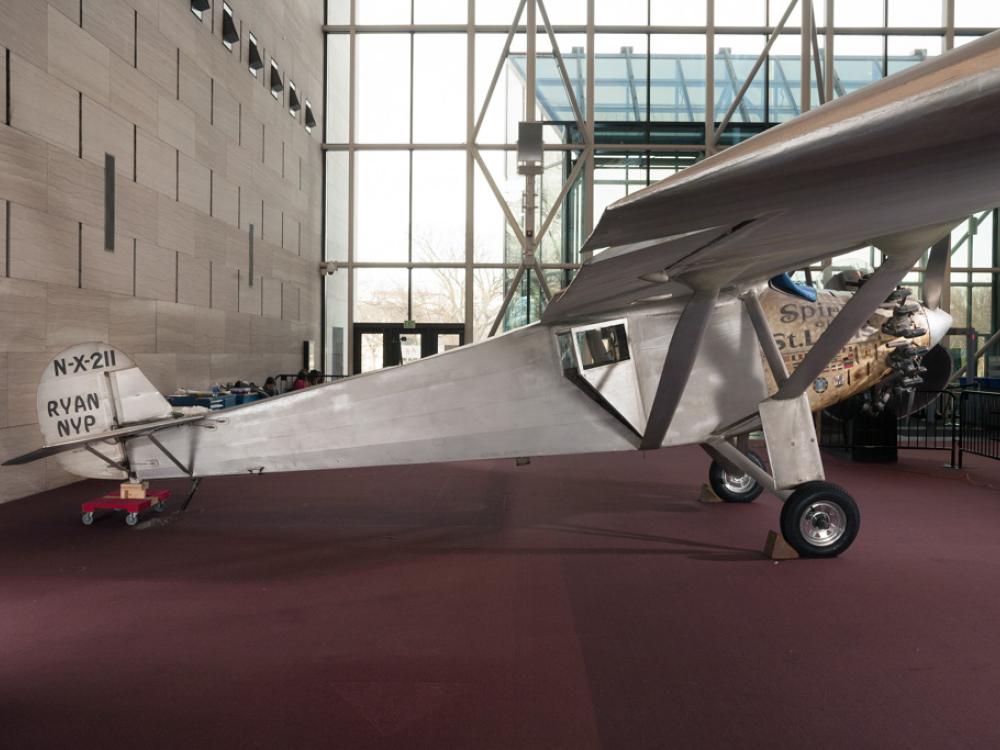 Side view of the Spirit of St. Louis, a silver-colored monoplane, on the floor of the Museum. Registration number "N-X-211" is painted on the tail above the make of the plane, "Ryan MVP".