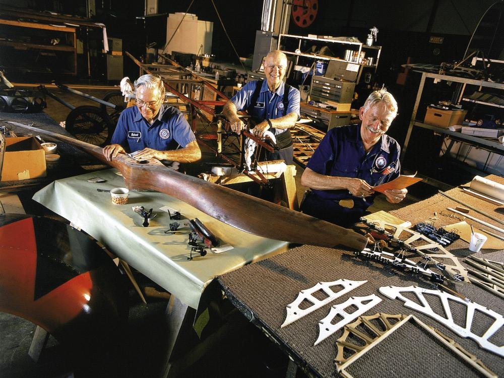 Three restoration volunteers are seated and smiling while working on aircraft parts. A volunteer in the foreground holds sandpaper over the center of a large wood propellor. 