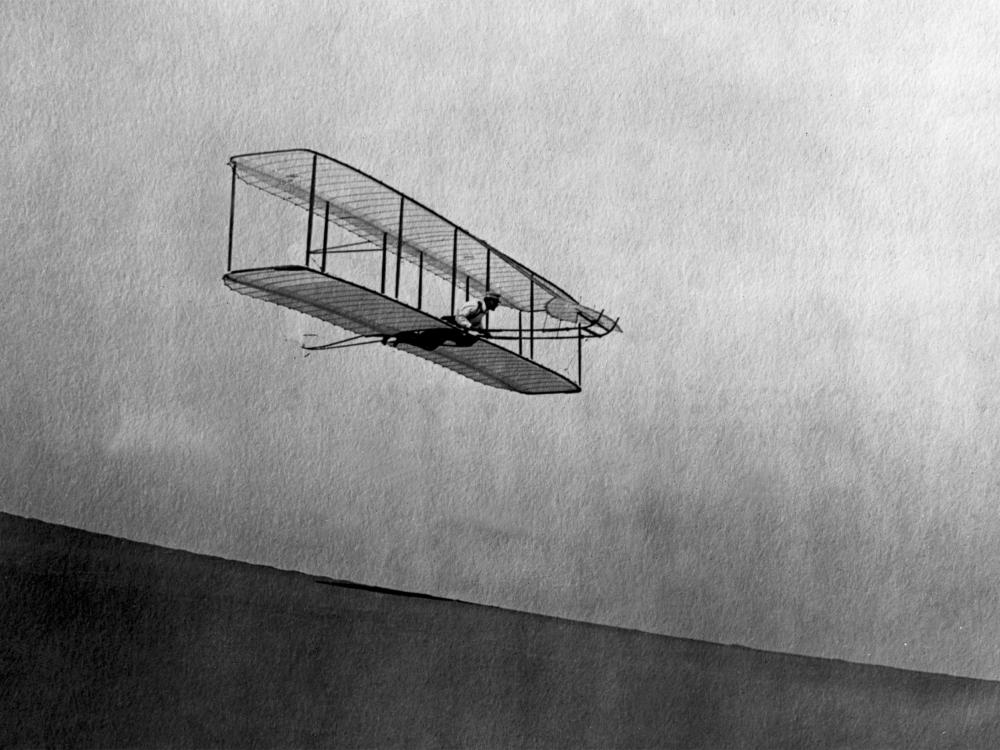 A biplane gliding in the air down a hill. A person lays on top of the bottom wing, perpendicular to it, as they pilot the craft.