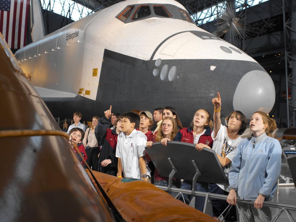 Students stand and point at artifacts hanging above with the Space Shuttle Enterprise in the background.