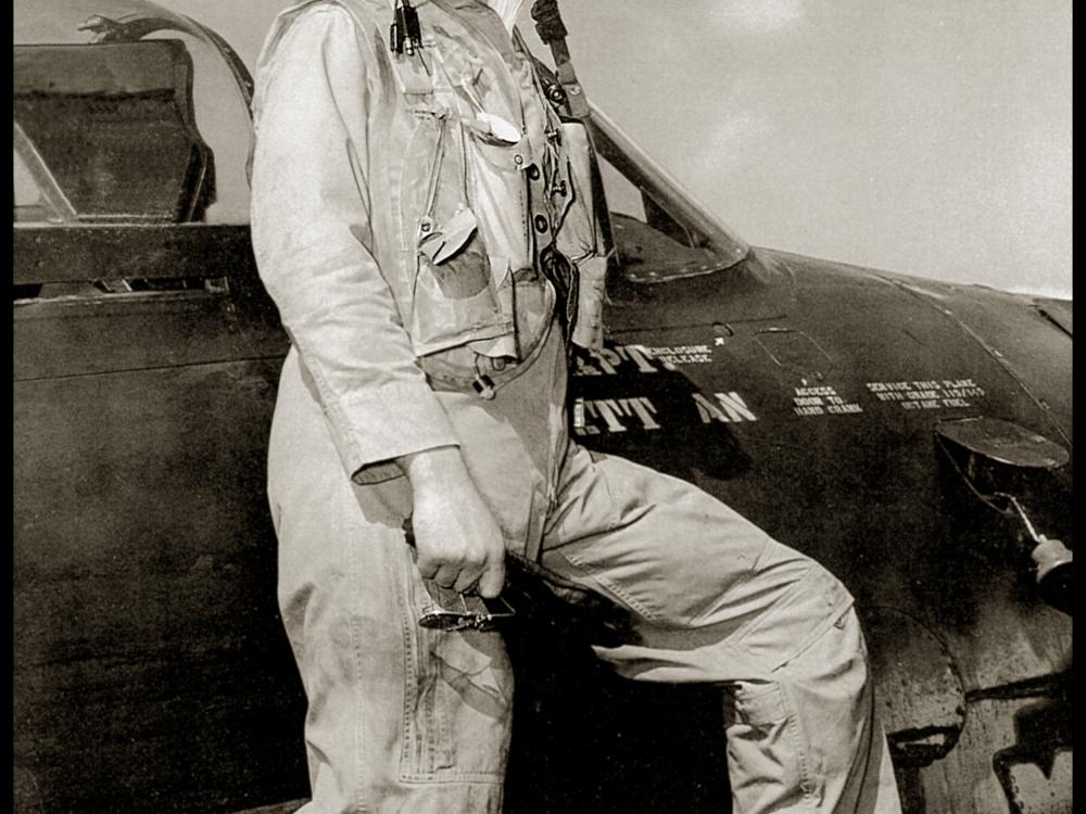 Bill Daniels, a man, stands on the wing of a Grumman F8F Bearcat while wearing aviator gear.