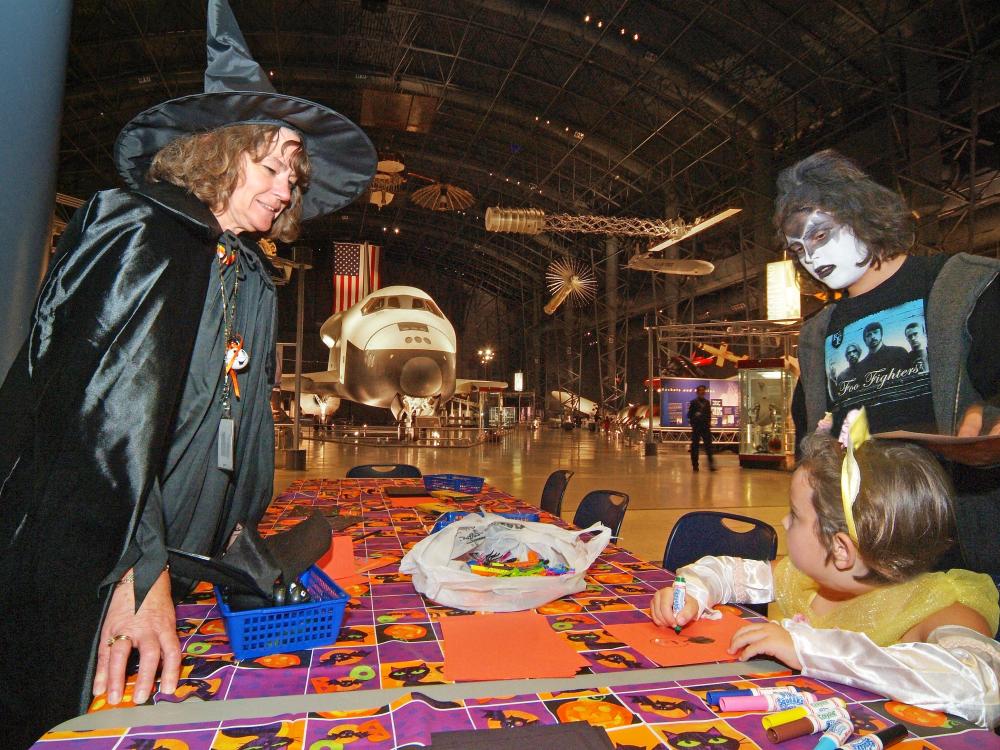 Trick-or-treater vistors stand at a Halloween-themed display with a museum curator dressed in a witch costume.