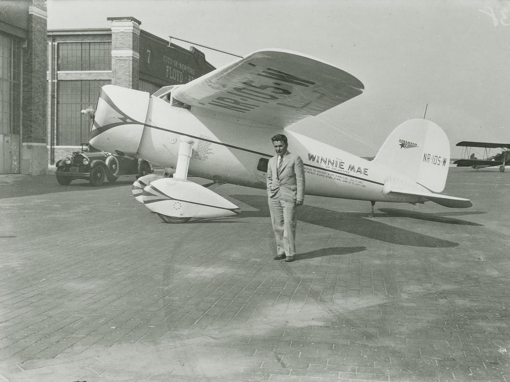 Wiley Post stands on the side of his Winnie Mae aircraft, under its left wing. 