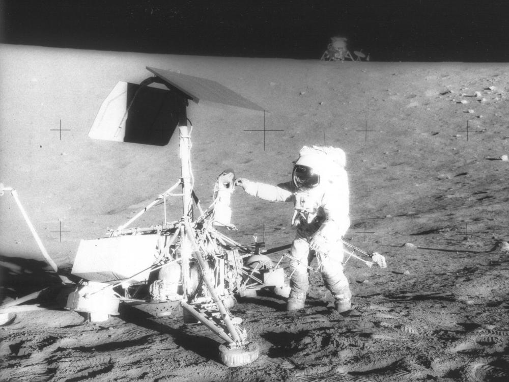 An astronaut on the moon during the Apollo 12 mission works next to and with a large camera set up with solar panels.