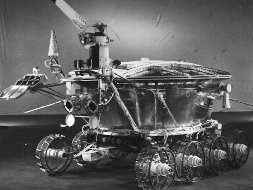 Black and white image of a robot lunar rover with a pie-shaped base and multiple probes, cameras, and antennae attached to the base.