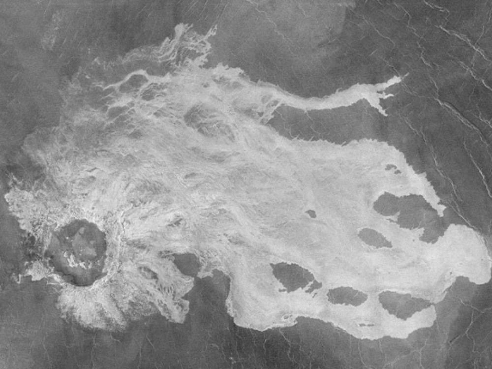 A black and white satellite view of a section of Venus's surface with a large crater impact and lava flows surrounding the crater. The lava flows are lighter in color than the rest of the visible surface.