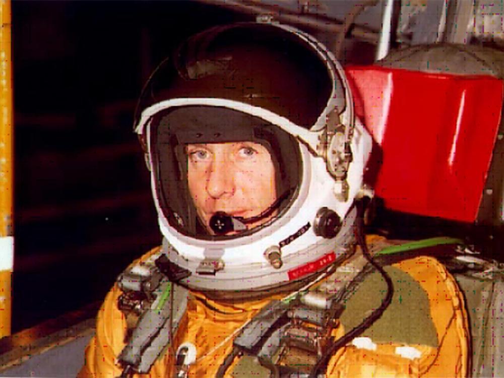 Col. Richard Graham, a white man, sits in an orange and green flight suit and white helmet.