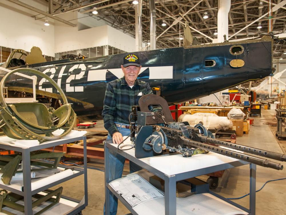 Charles French, a white male veteran, poses in the Restoration Hangar of the museum in front of the Helldiver.