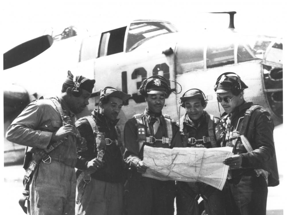 Five Black aviators look at a map. They're dressed in flight gear. There is a plane behind them.