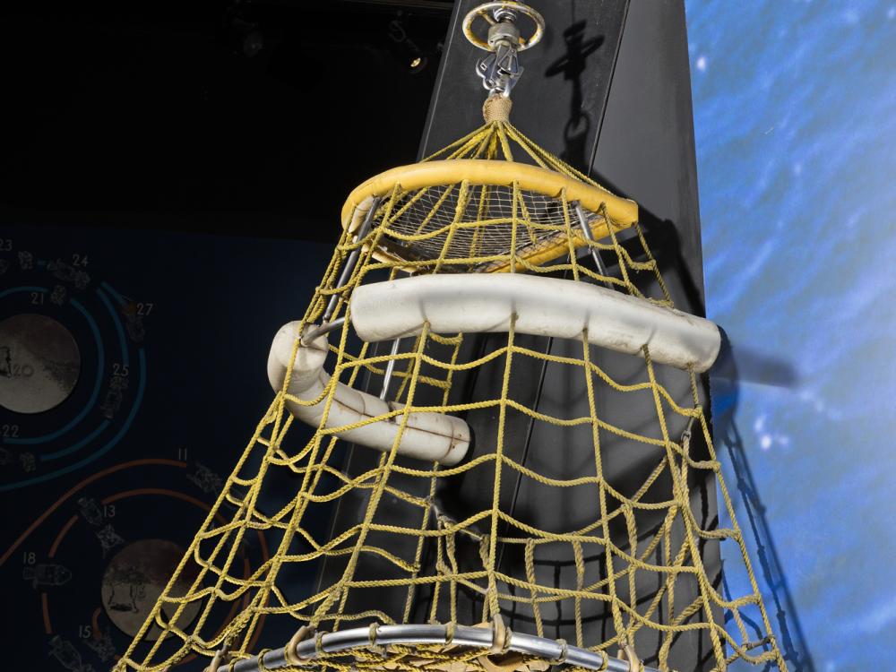 Quadrilateral-shaped metal frame with yellow fabric cords and two white pontoons near top hanging in musuem