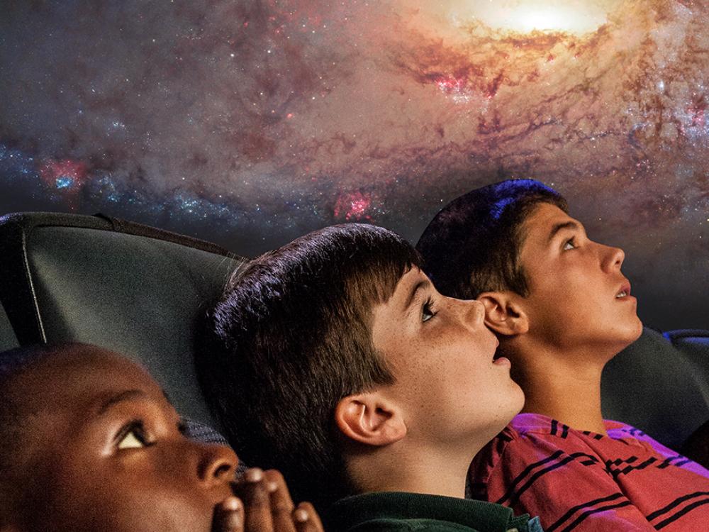 Three boys look up at a planetarium show that is above their heads. 