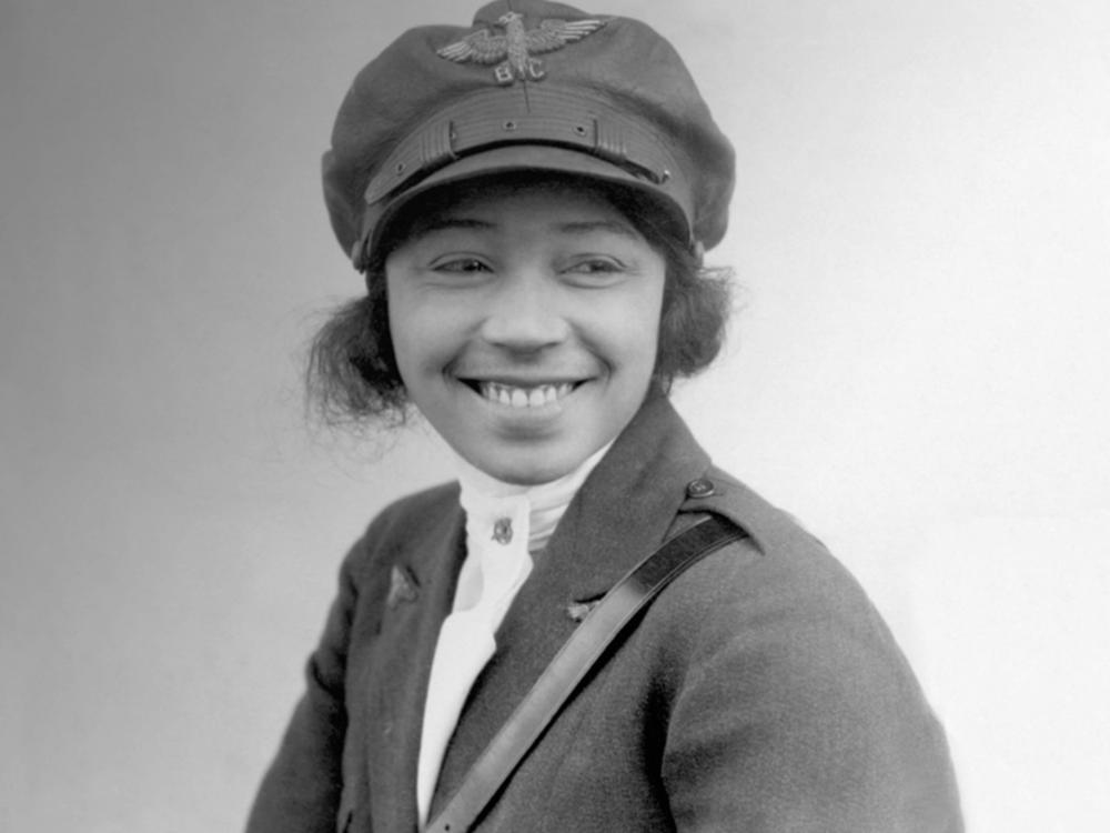 Bessie Coleman, a woman who appears to be in her late 20s, smiles at something to the right of the camera. She is wearing a military-like uniform, with an emblem that reads "B.C." on her cap.