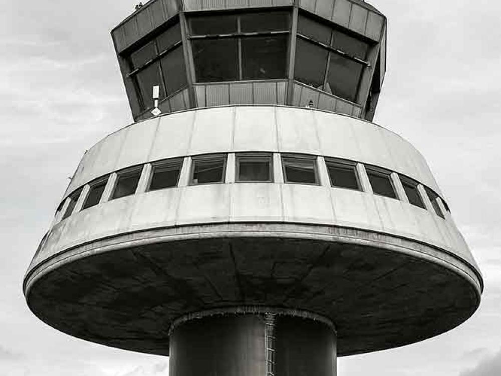 Partial view of an air traffic control tower at Barcelona El-Prat Airport. This tower features a dark-colored cylinder base with a conical-shaped first floor. A second floor features at least four different perspectives for observation.