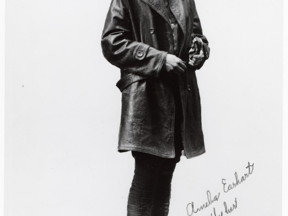 Amelia Earhart, a woman, poses for a photo in aviator gear. Her name is written in cursive script next to her legs.