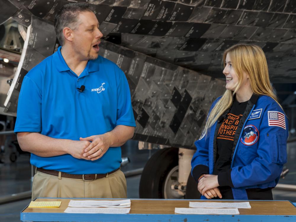 Man in blue shirt talks to woman in astronaut jacket