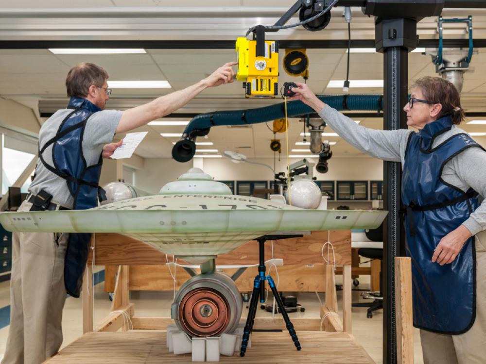 Two Smithsonian employees position an X-ray scanner on top of a large, dish-shaped spaceship studio model.