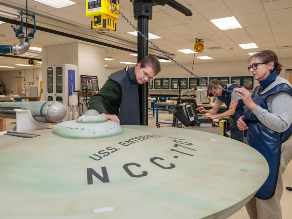 Three Smithsonian staff members use an X-ray machine to take images of the interior of a dish-shaped spaceship studio model.